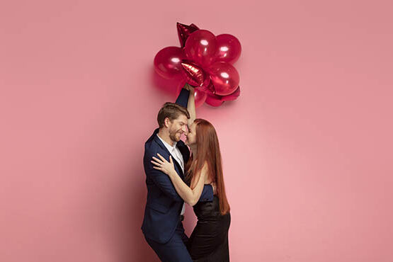 Happy holding balloons shaped hearts. Valentine's day celebration, happy caucasian couple on coral background.